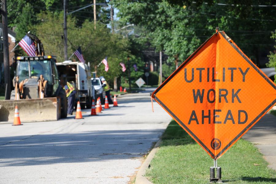 An orange diamond-shaped sign saying Utility Work Ahead sits on a street in front of construction equipment and workers