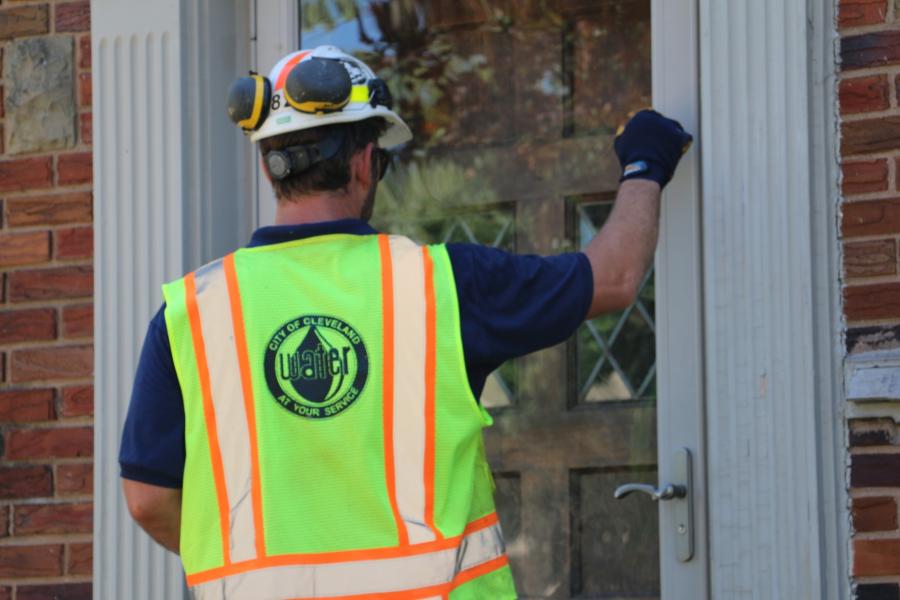 A man wearing a hard hat and yellow high-visibility vest with a Cleveland Water logo on the back knocks on the front door of a house.