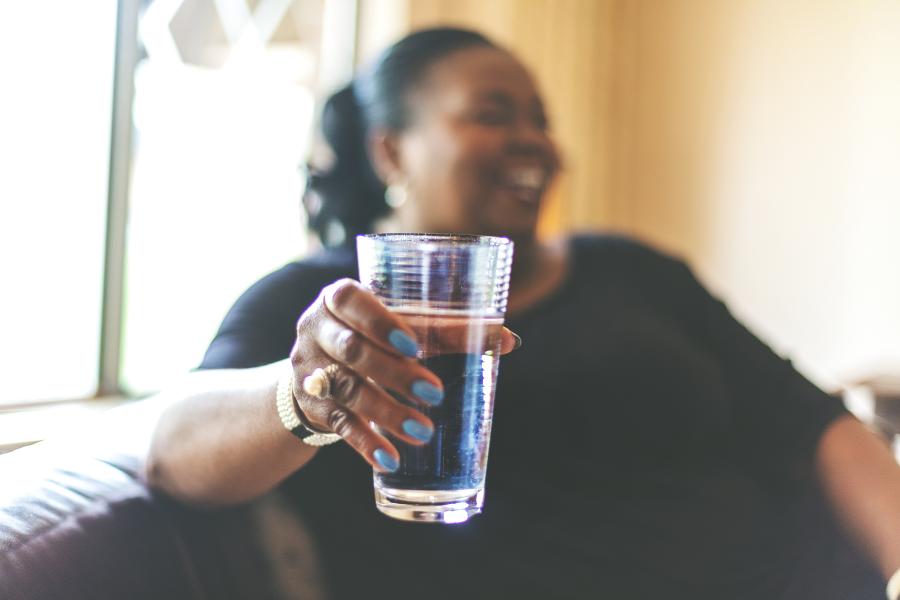 A woman sits on a couch holding a glass of water and smiling