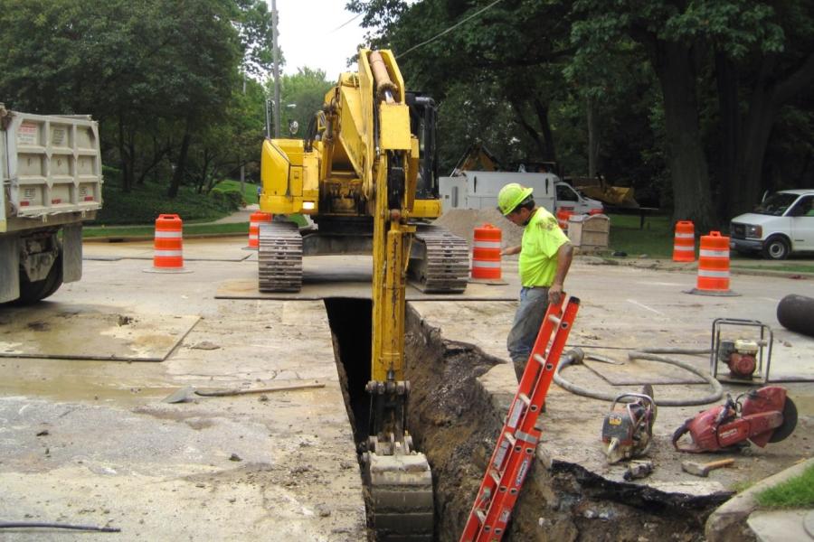 An excavator digs a trench in a road for a new water main while a crew member looks on.