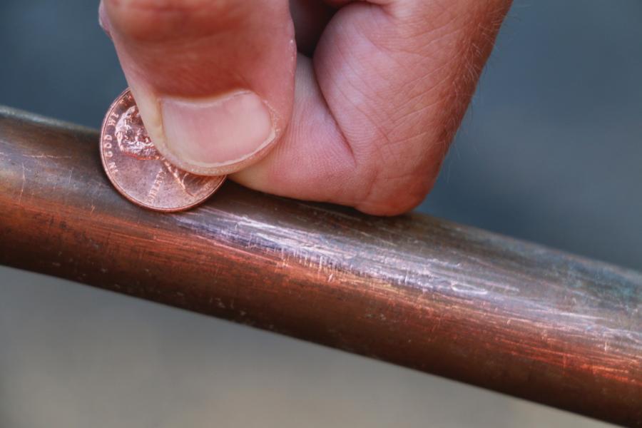 A man's hand scratching a copper pipe with a penny