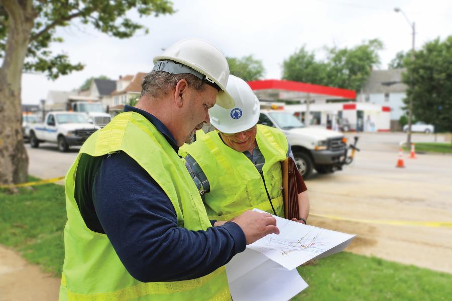 Two men wearing hard hats and yellow high visibility vests stand on a sidewalk next to a work zone looking at infrastructure maps