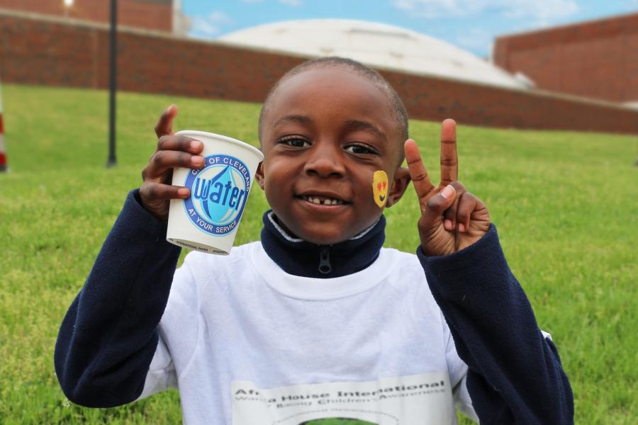 A smiling boy with a smiley face painted on his cheek holds up a Cleveland Water cup and gives the peace sign