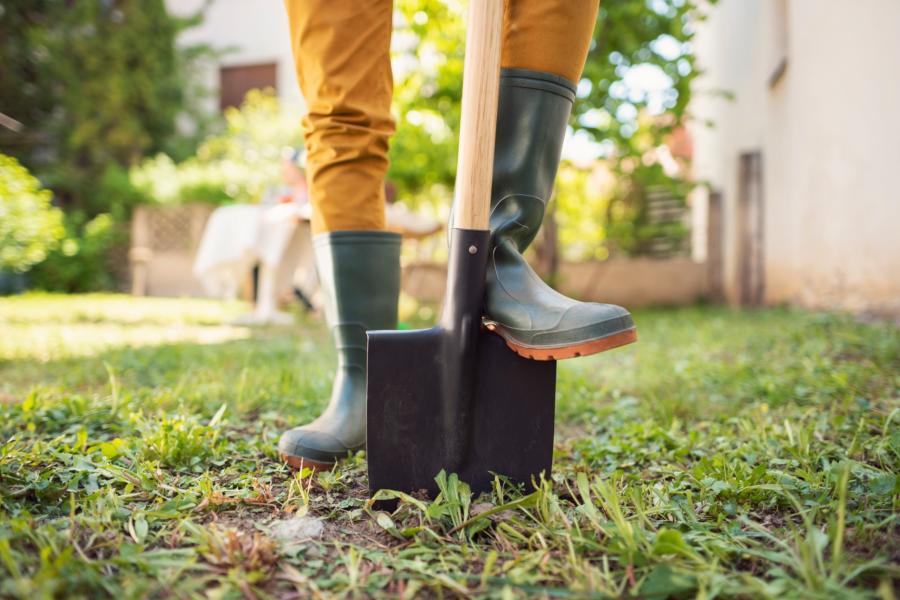 A close up of someone wearing green rain boots and yellow pants with one foot on a shovel digging into a yard