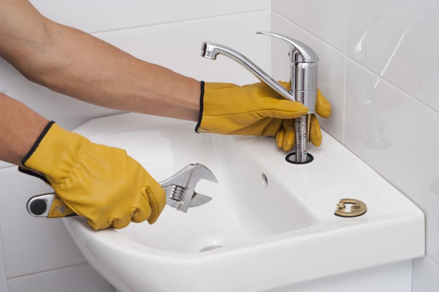 A man wearing yellow work gloves and holding a wrench in one hand works on a bathroom faucet.
