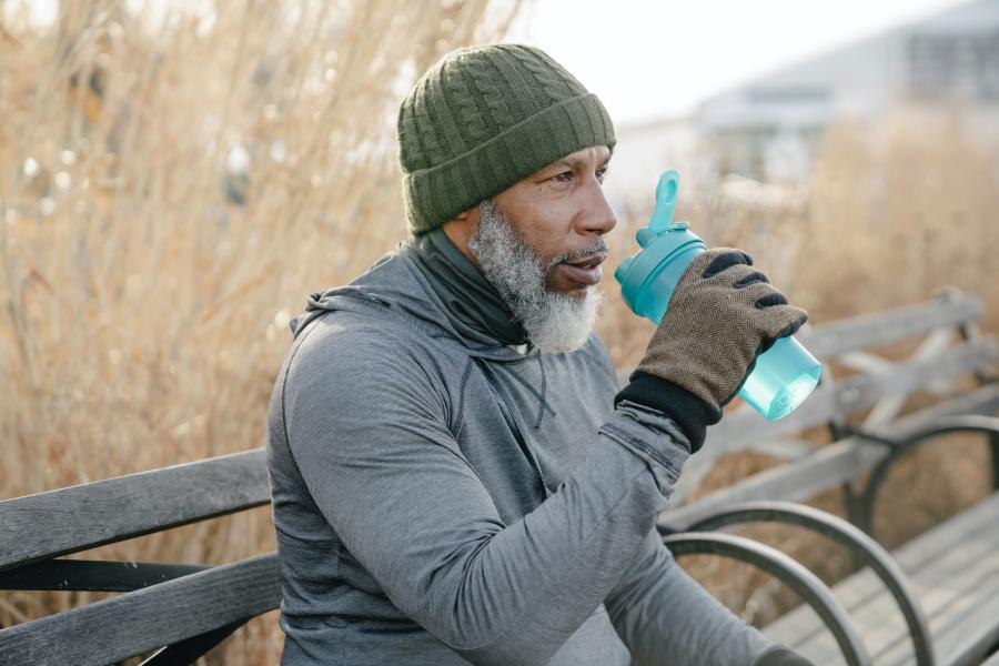 A man dressed for cold weather sits on a bench drinking water from a water bottle
