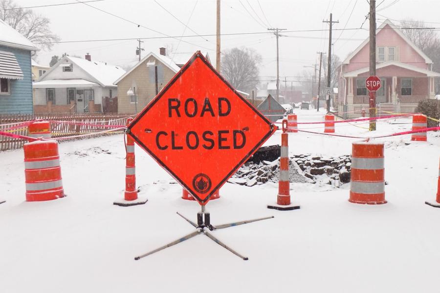 An orange diamond-shaped sign saying Road Closed sits on a snow-covered street in front of orange construction barrels and cones