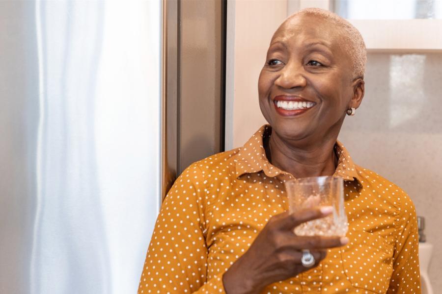 An older black woman stands in her kitchen smiling and holding a glass of water