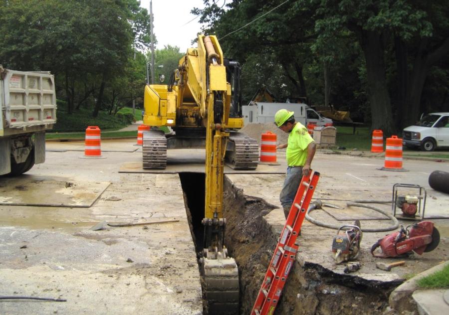 An excavator digs a trench in a road for a new water main while a crew member looks on.