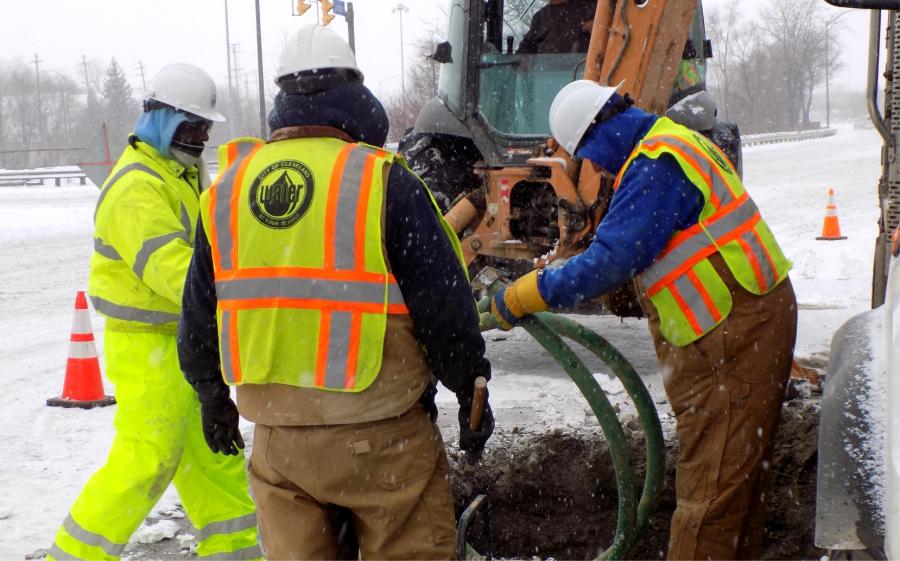 Three men dressed in construction gear and winter clothes surround an excavation in a roadway with construction equipment in the background