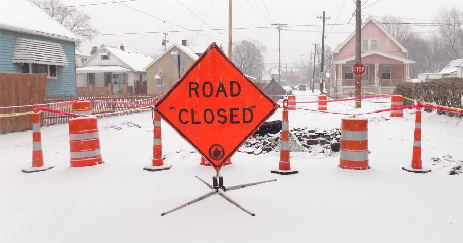 An orange diamond-shaped sign saying Road Closed sits on a snow-covered street in front of orange construction barrels and cones