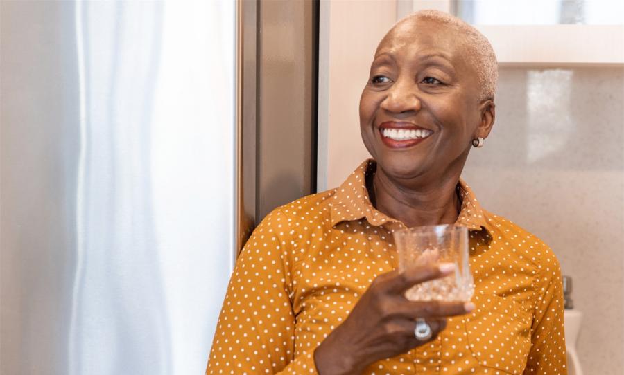 An older black woman stands in her kitchen smiling and holding a glass of water